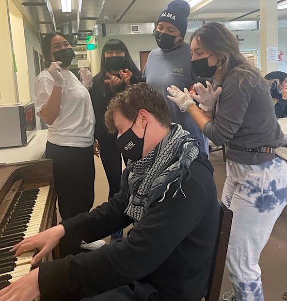 Student playing the piano with other students gathered around