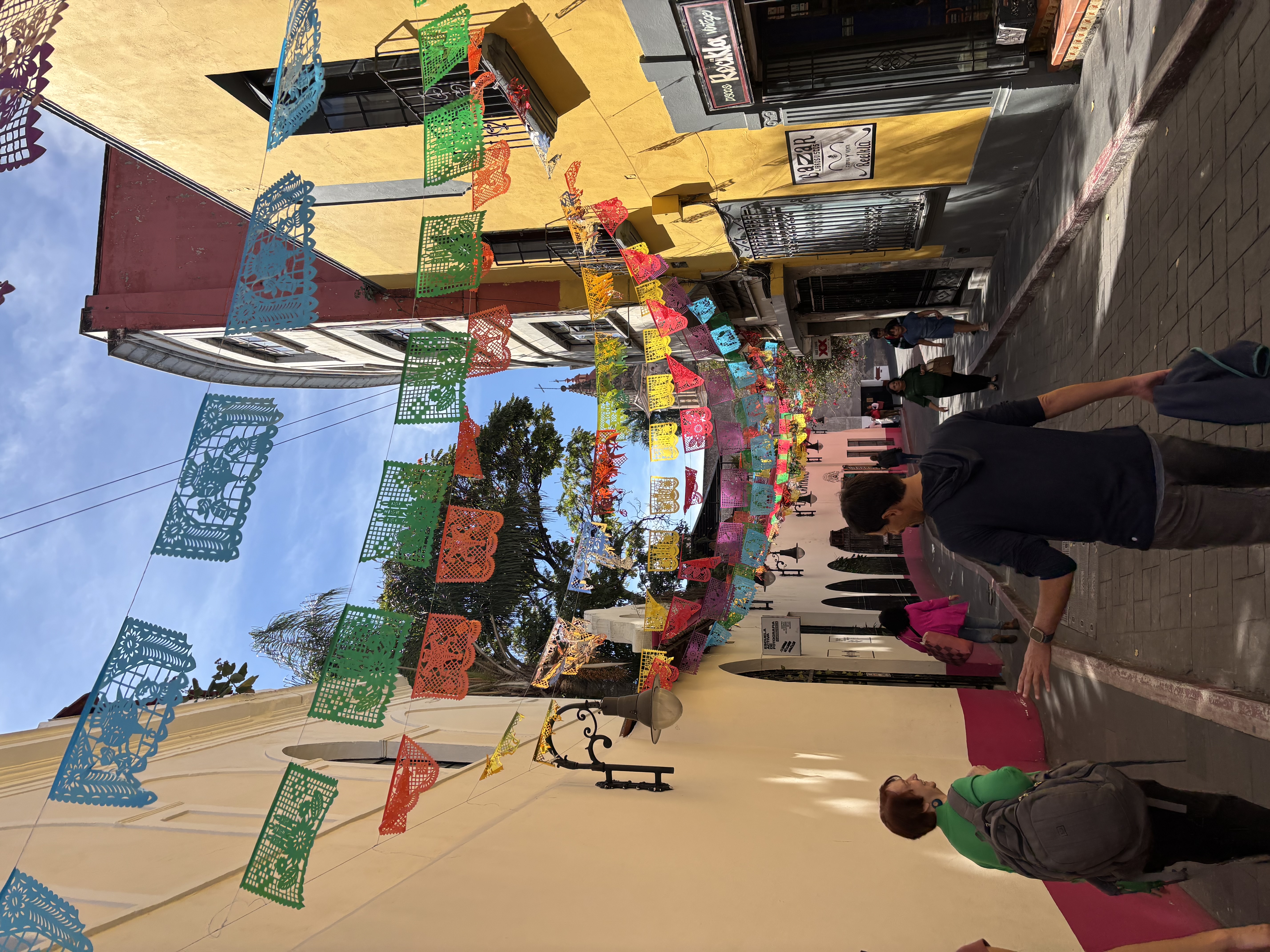 A group of people walk along colorful streets in Cuernavaca, with papel picado garlands above them