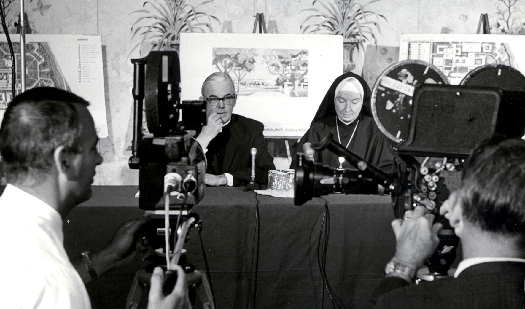 A vintage image of a press conference showing a priest and a nun at a table signing documents with a camera crew filming them